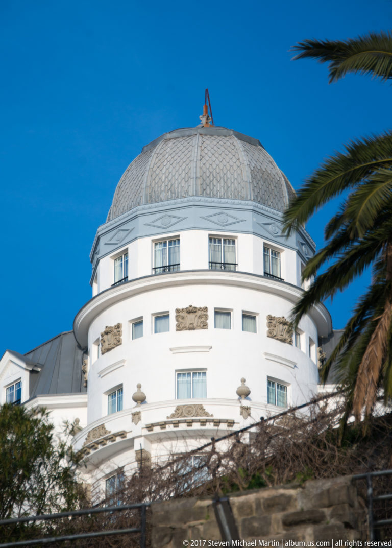Round white building with rectangular windows and a broad faceted metal dome, partly framed by palm fronds.
