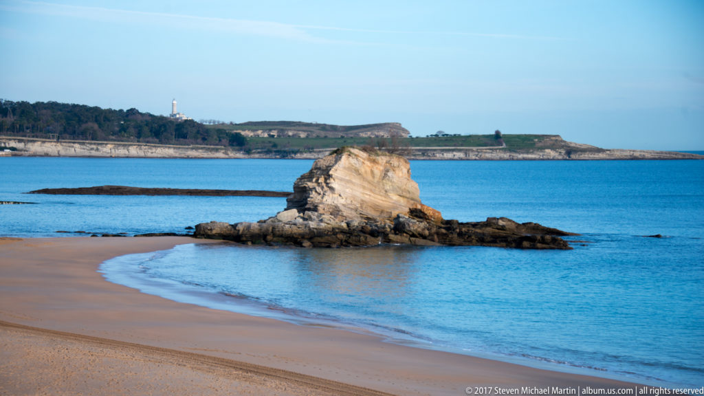 Eroded rock stack just offshore from a sandy beach, with blue water and a wooded headland in the distance.