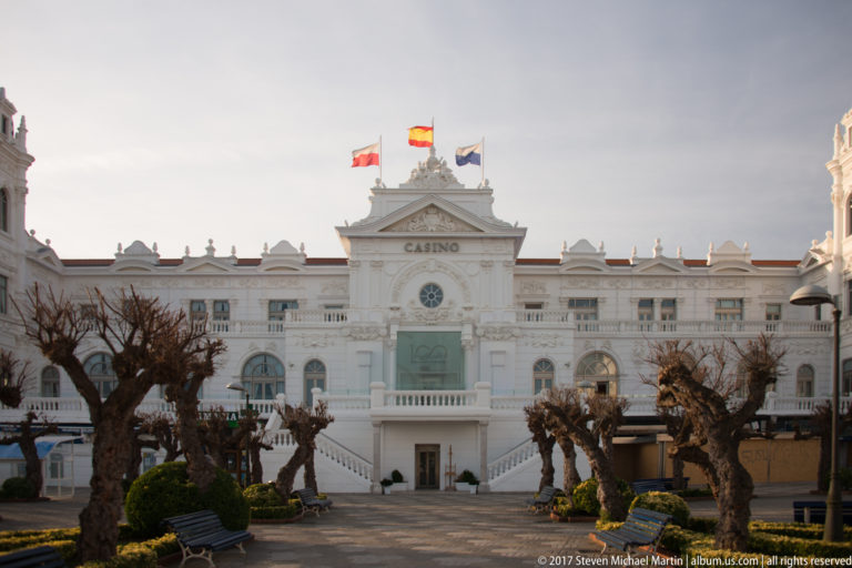 Frontal white façade of Gran Casino del Sardinero with central pediment, clock, flags, and pollarded trees.