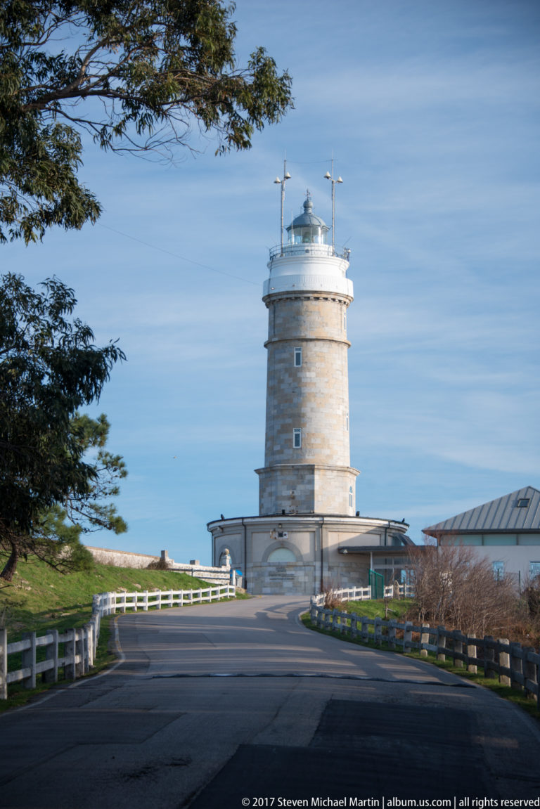 Cylindrical stone Cabo Mayor Lighthouse beside a curving access road, with white lantern gallery and low service buildings.