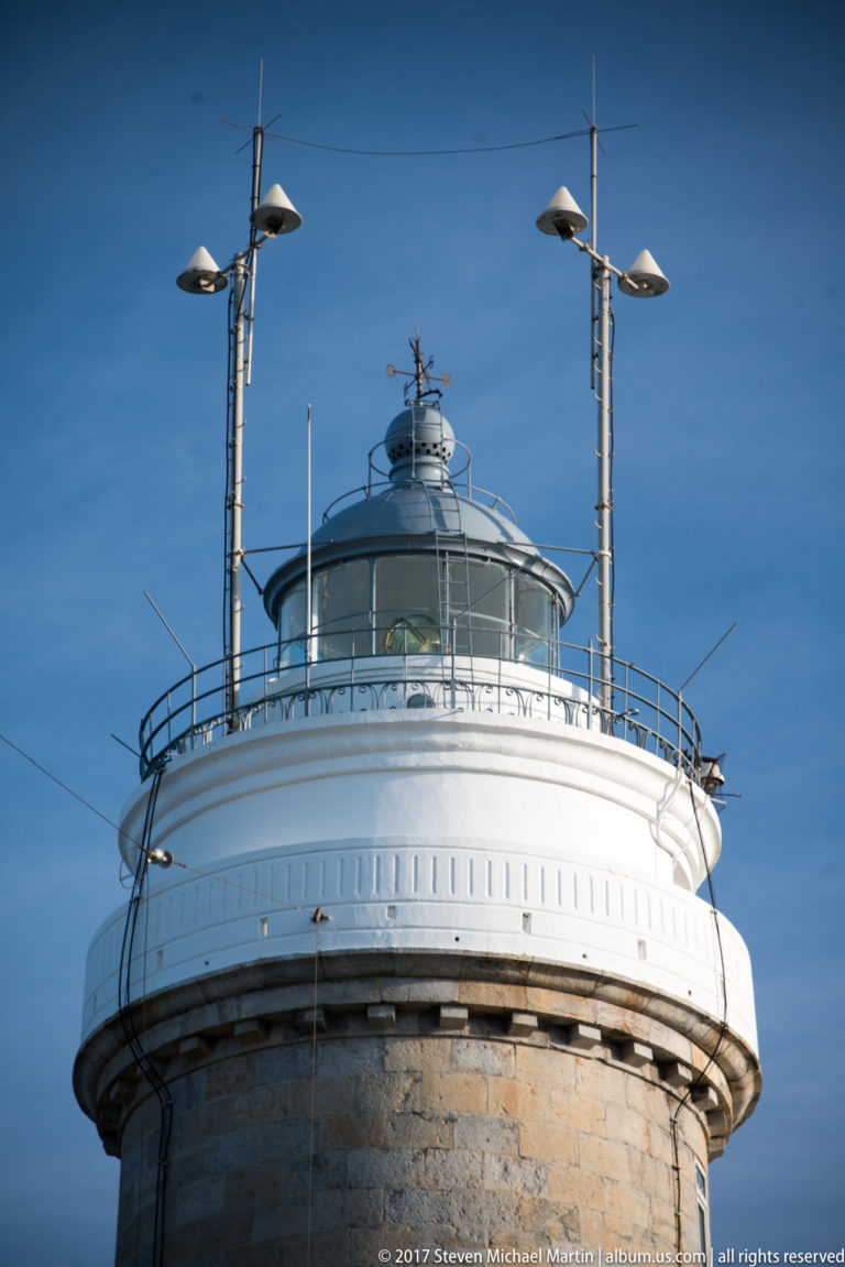 Close view of Cabo Mayor Lighthouse upper tower, white gallery, glazed lantern room, railings, antennae, and fixtures.