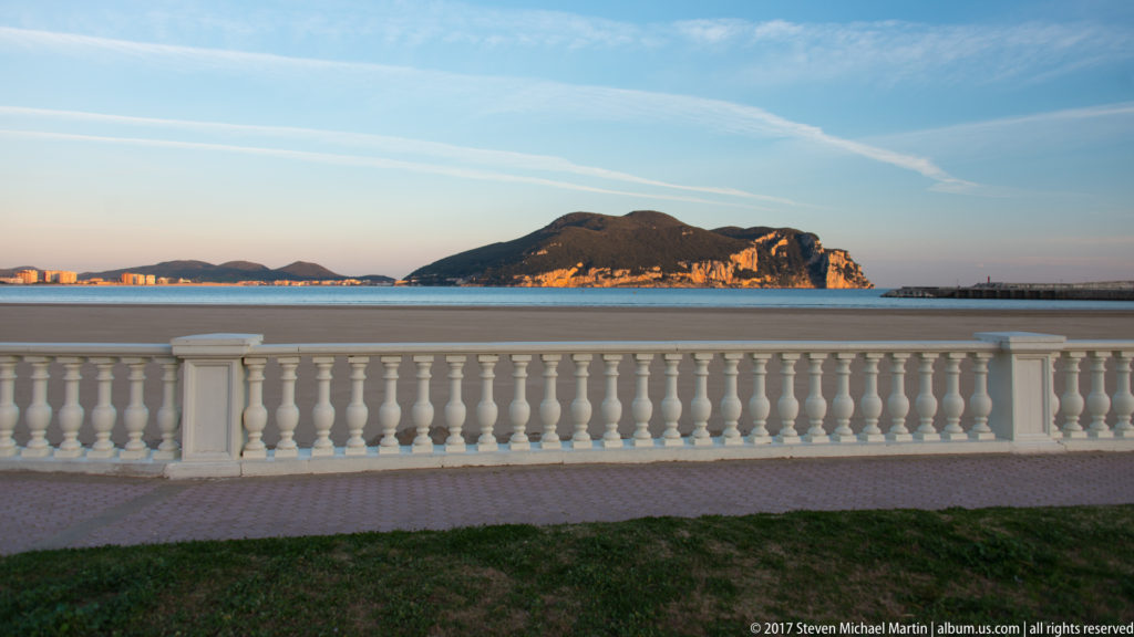 White balustrade along a waterfront promenade with a rocky island across blue water in warm light.
