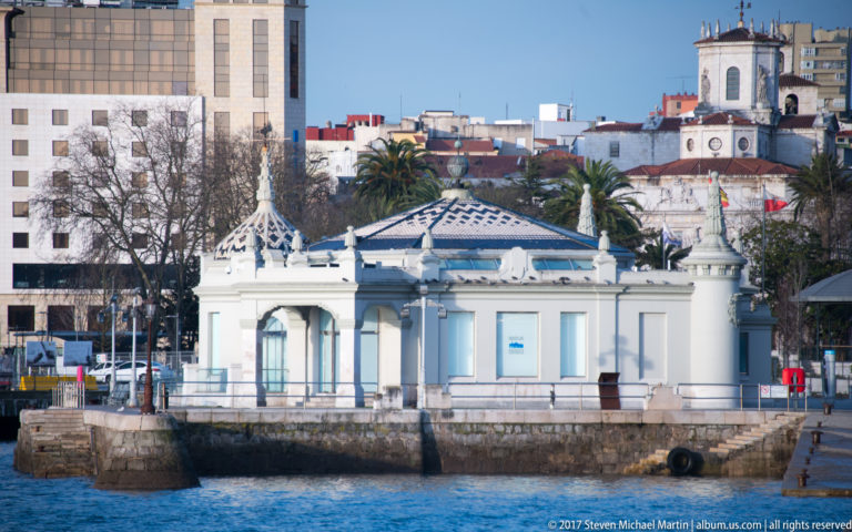 Pale waterfront pavilion on a stone quay, with decorative roof details, palms, and city buildings behind.