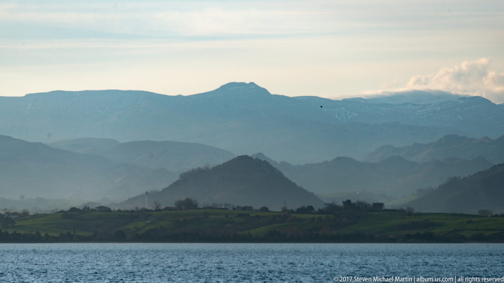 Calm bay water before green shore and layered blue-gray mountains, with higher partly snowy peaks in the distance.
