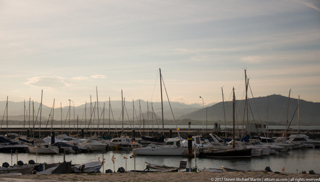 Rows of moored sailboats and tall masts in a marina, backed by hazy hills and mountains.