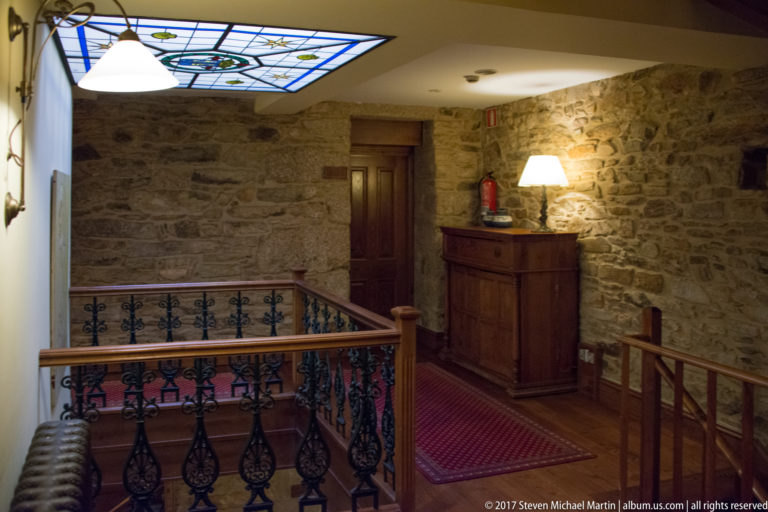 Stone-walled interior landing with iron railing, wooden floor, red runner, cabinet, and skylight.