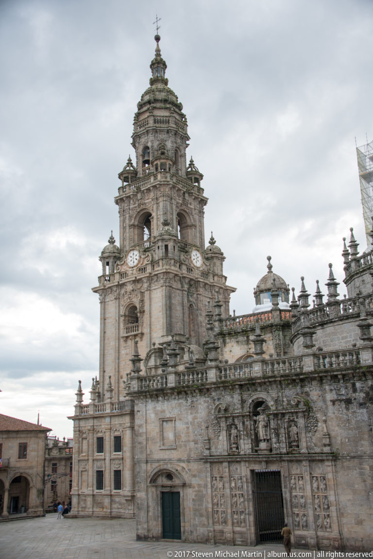 Oblique plaza view of a cathedral tower joined to a long sculpted stone façade with balustrades and rooftop finials.