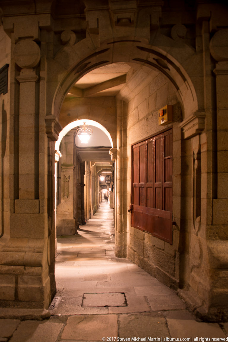 Warmly lit granite arcade with repeating arches, stone columns, wooden shutters, and a long pedestrian passage at night.