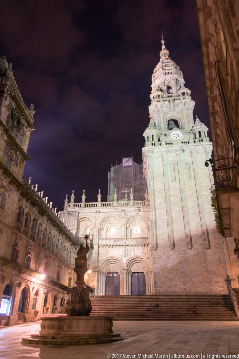 Illuminated stone cathedral tower and adjoining façade at night, seen across steps and a fountain.