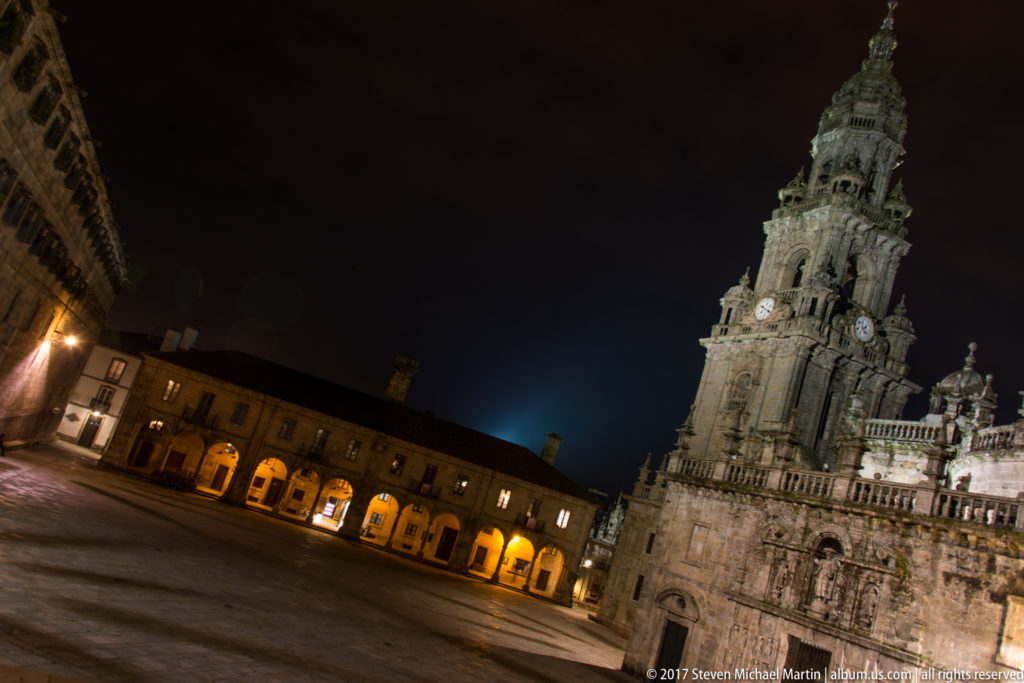 Tilted nighttime view of an illuminated cathedral clock tower across a broad plaza lined by warm-lit arcades.