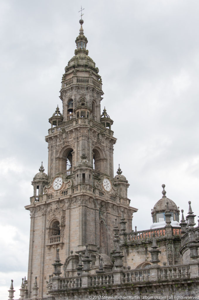 Ornate cathedral clock tower rising above a balustraded roofline and smaller dome under a gray sky.
