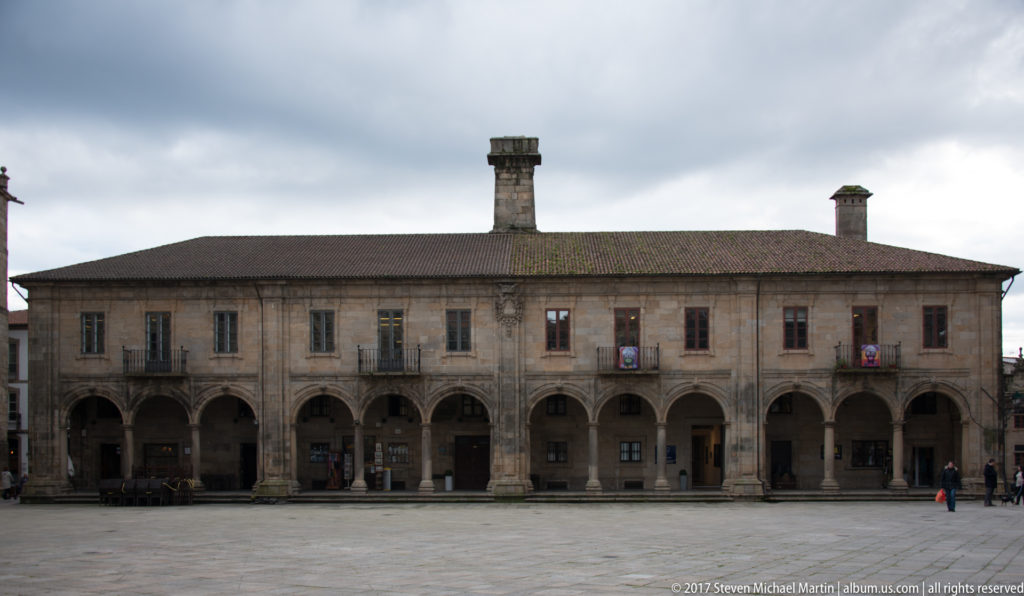 Long two-story granite building with repeated ground-floor arches, upper windows, hipped roof, and central chimney.