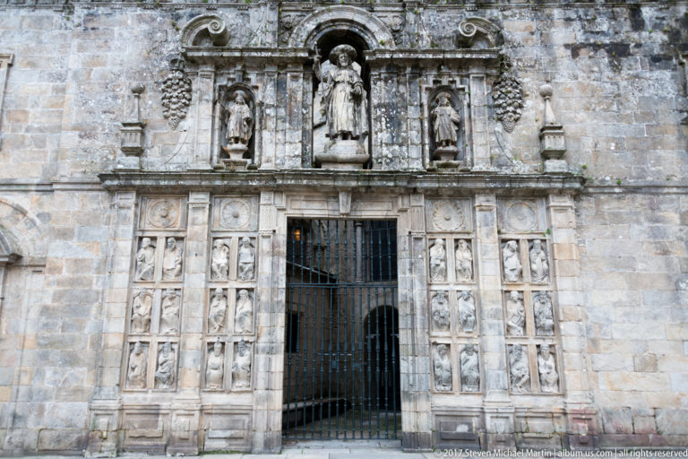 Iron-gated cathedral entrance framed by rows of carved relief panels and saint statues in pale weathered stone.
