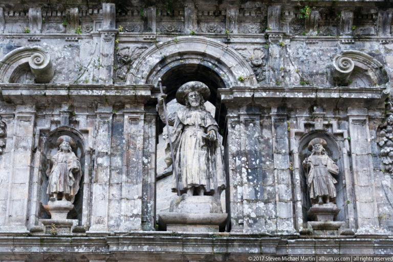 Three stone saint statues in exterior niches, with a larger staff-bearing pilgrim figure centered beneath a rounded arch.