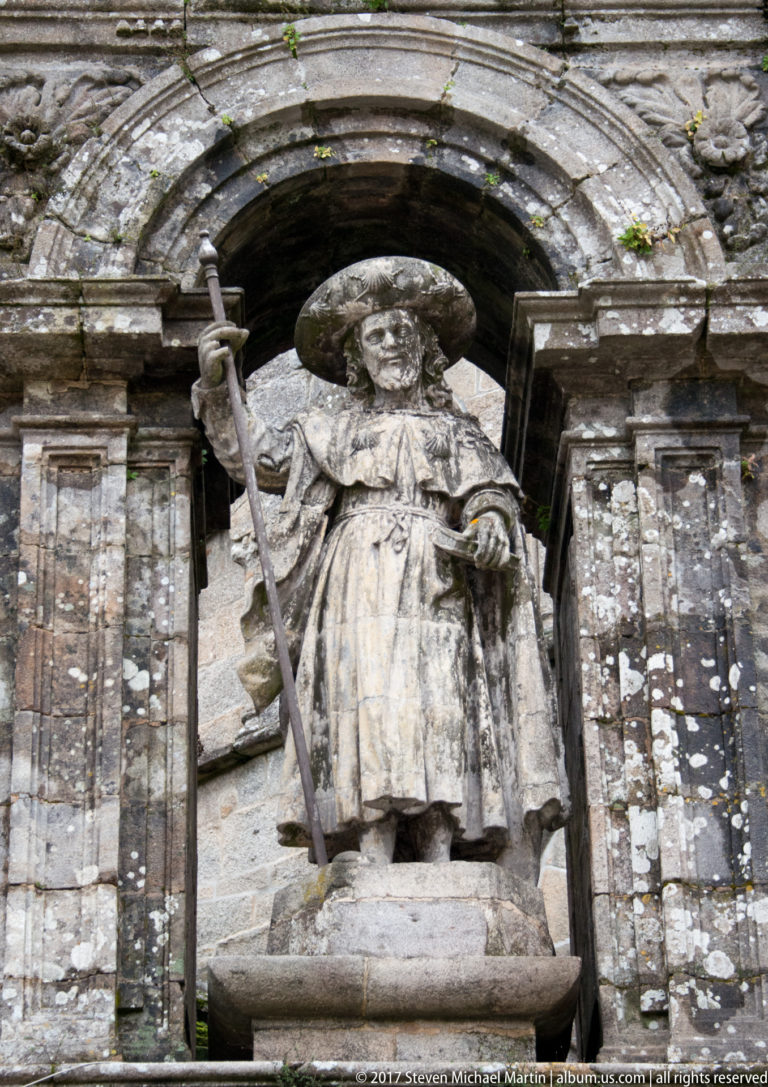Weathered stone pilgrim saint holding a long staff beneath a shell-like canopy in a dark architectural niche.