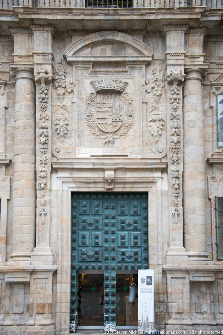 Frontal stone cathedral doorway with blue-green paneled doors, paired columns, carved ornament, and a heraldic panel above.