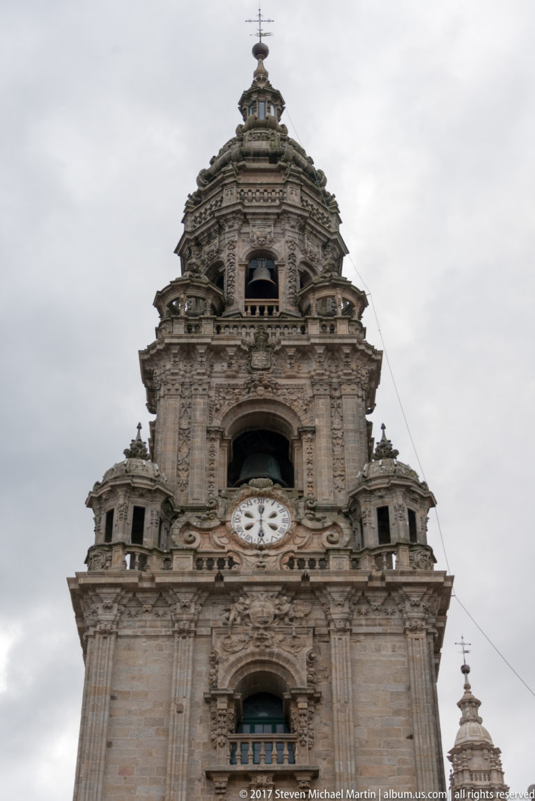 Upward view of an ornate stone cathedral tower with clock face, open belfry, pinnacles, and crowning lantern.