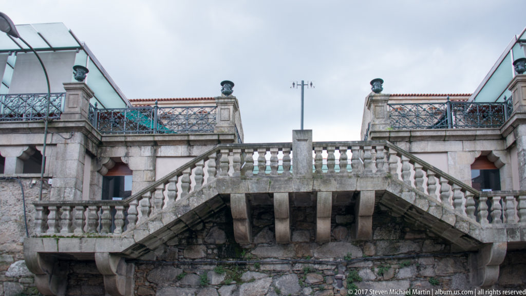 Symmetrical stone staircase with balustrades, projecting landing, finials, and metal-railed terraces under a cloudy sky.