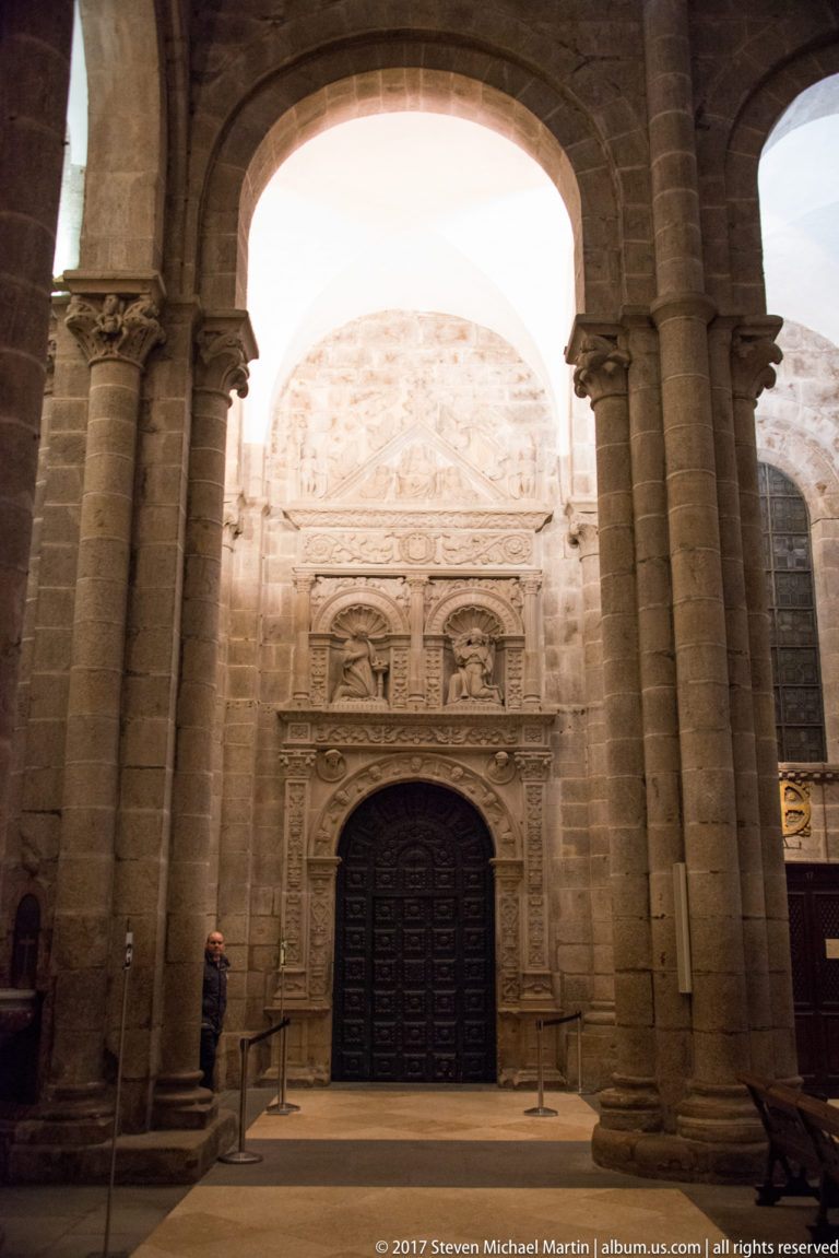 Stone columns frame a richly carved portal with dark double doors inside a vaulted cathedral passage.