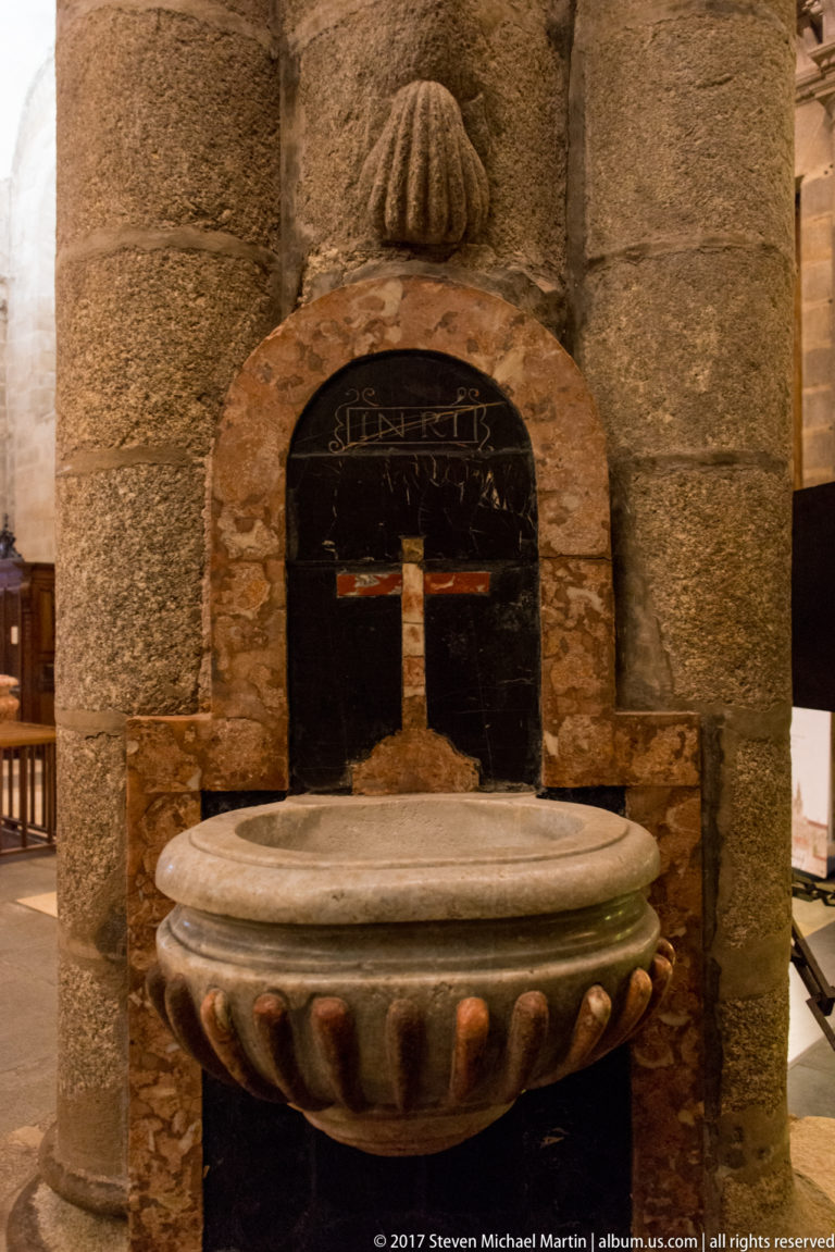 Stone basin on a granite wall beneath a carved scallop shell, with a dark inset panel and pale cross behind it.