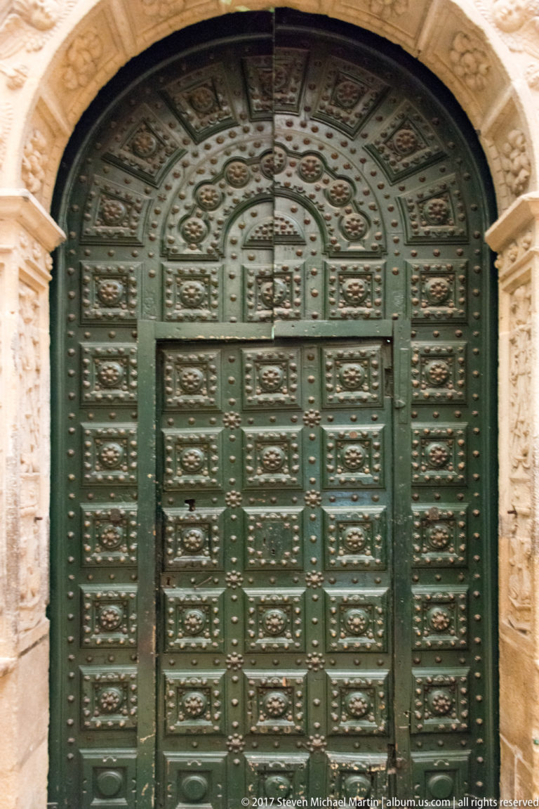 Large green arched cathedral door divided into rectangular relief panels with metal studs, framed by pale carved stone.