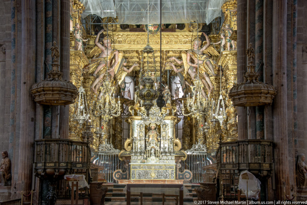 Densely gilded cathedral sanctuary and altar framed by massive dark stone piers and hanging lamps.
