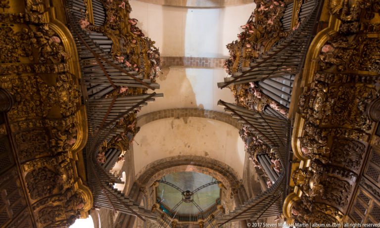 Upward view between two richly carved organ cases with dark pipes, gilded ornament, and the cathedral vault between them.