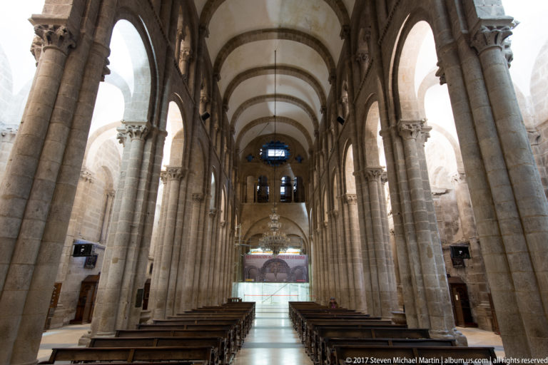 Tall Romanesque cathedral nave with rounded arches, massive stone piers, wooden pews, and a bright central aisle.