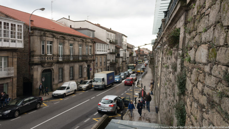 Cars and pedestrians on a narrow street between a massive granite wall and historic buildings with glazed galleries.