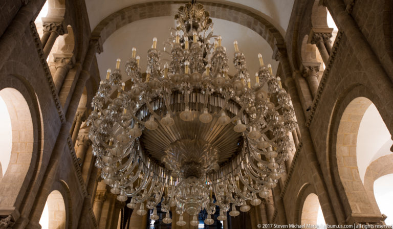 Close view of a huge multi-tiered crystal chandelier suspended among rounded stone arches and columns.