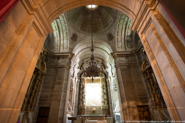 Stone arch framing a vaulted cathedral bay with a hanging chandelier and bright altar before a window.