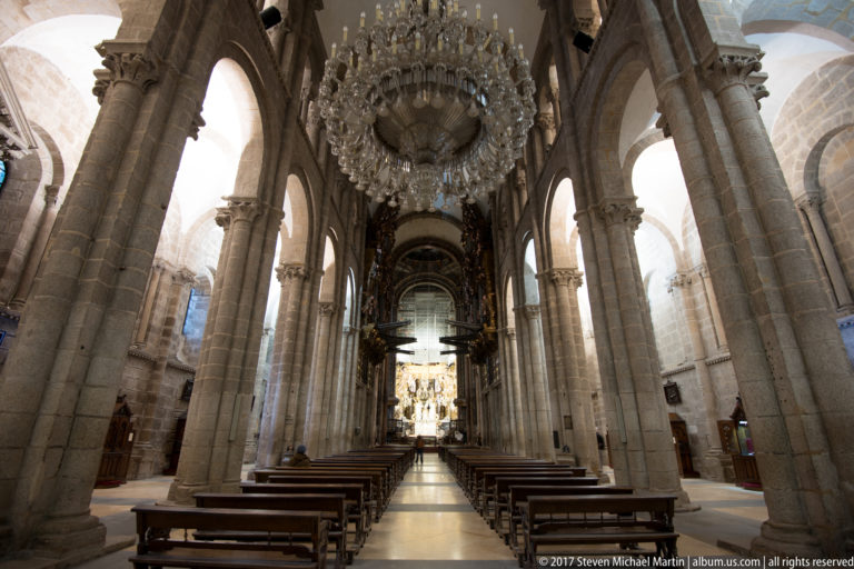 Long cathedral nave with wooden pews, massive stone piers, a huge glass chandelier, and a bright sanctuary beyond.