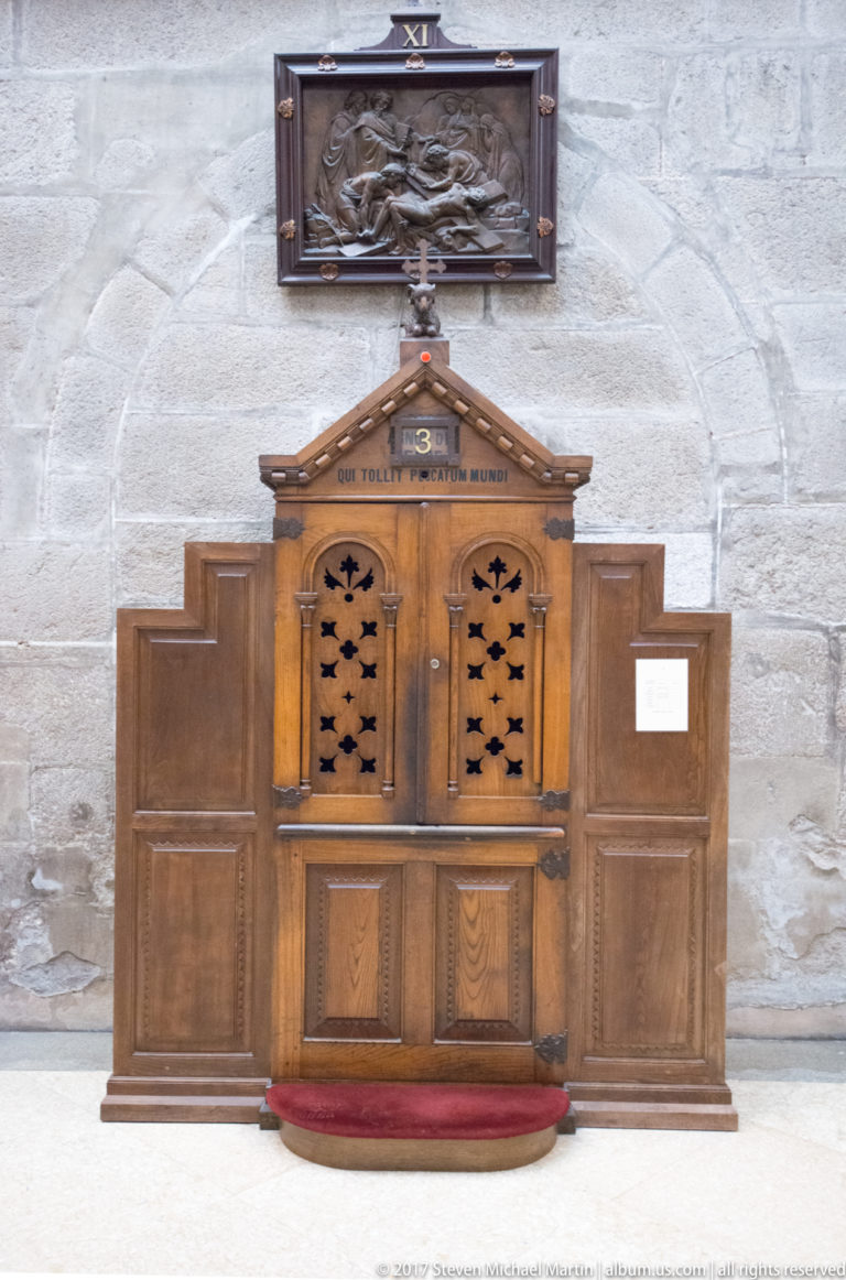 Wooden confessional with two screened openings and star-shaped cutouts, mounted below a dark carved relief on a stone wall.