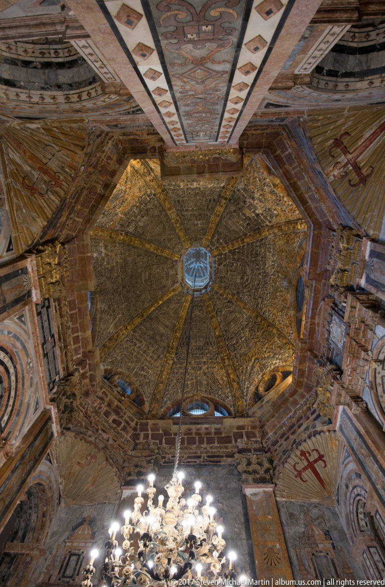 Upward view of an octagonal painted and gilded vault with geometric panels and a chandelier at the lower edge.