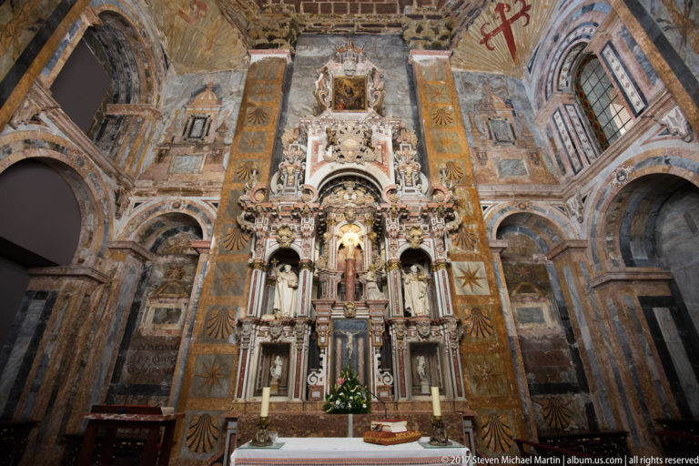 Tall multi-tiered altarpiece with religious figures, columns, flowers, and surrounding painted walls and arches.