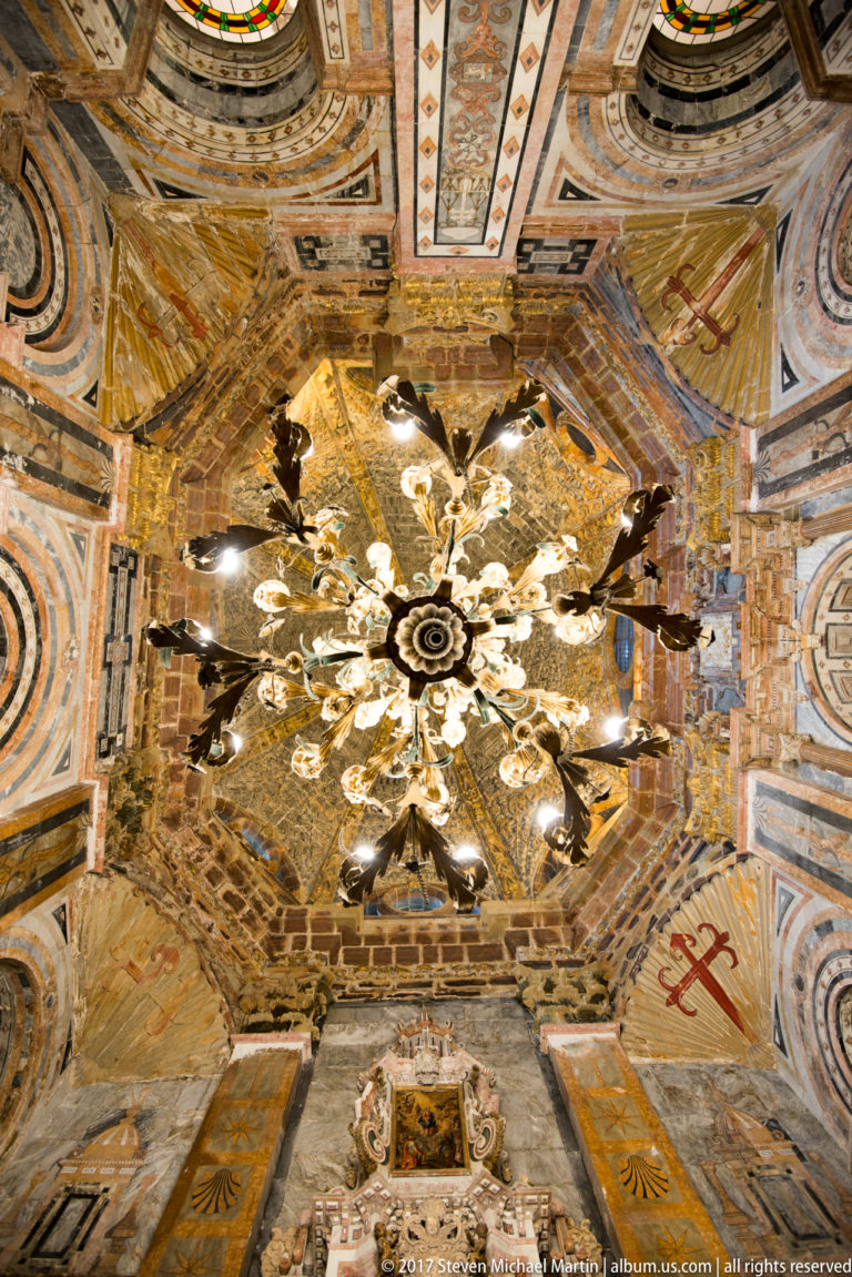 Large chandelier hanging beneath a richly painted and gilded vault, with decorated walls and an altarpiece edge below.