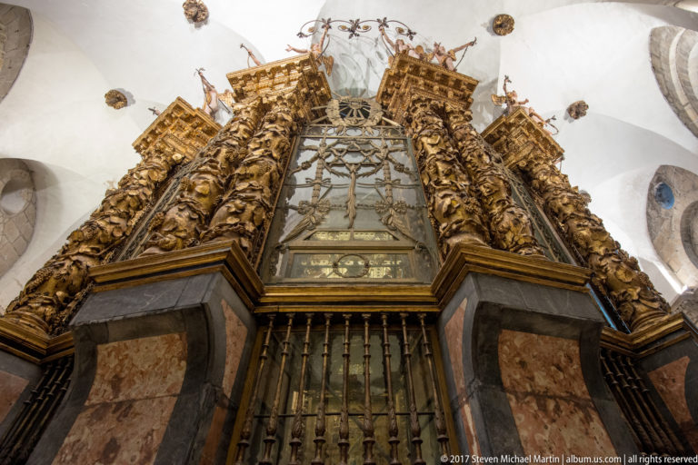 Low-angle view of a towering gilded carved structure with figures, foliage, projecting cornices, and a central glazed panel.