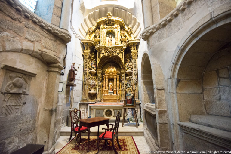 Small stone chapel with a gilded altarpiece, religious figure, table, and red-upholstered chairs.