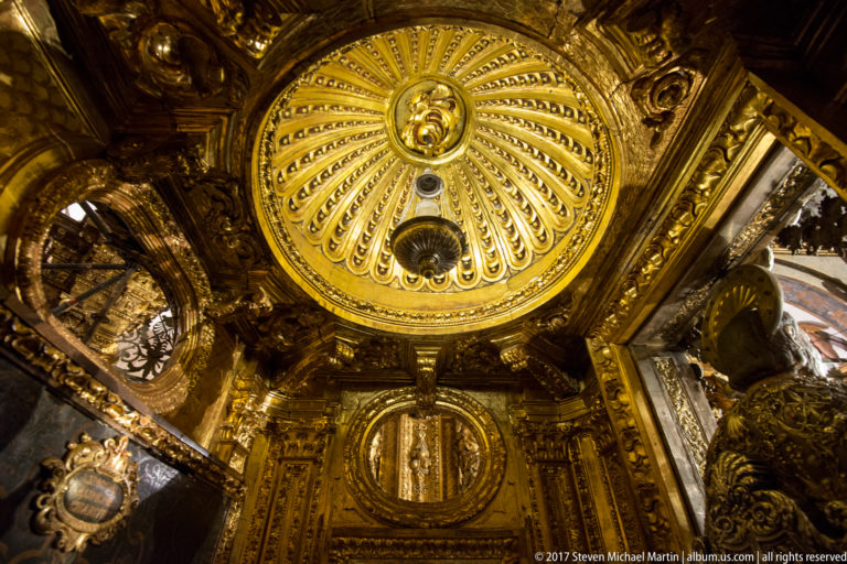 Upward view of a radial gilded ceiling with central medallion, hanging lamp, and ornate oval feature below.
