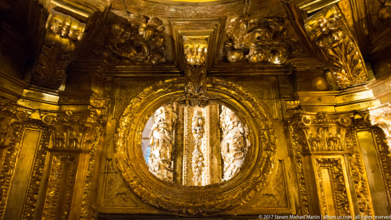 Close view of an oval opening surrounded by dense gilded carving, scrollwork, and sculptural ornament.