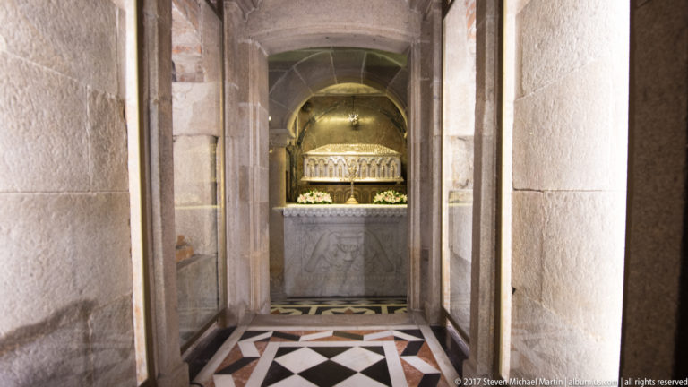Narrow stone passage ending at a small altar with flowers, beneath an arch and beyond a geometric floor.