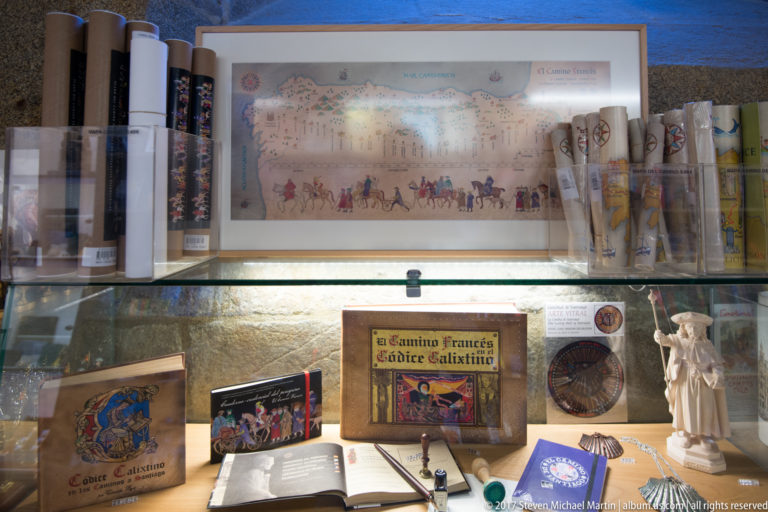 Display case containing Camino de Santiago guidebooks, maps, scallop-shell souvenirs, small objects, and a pilgrim figurine.