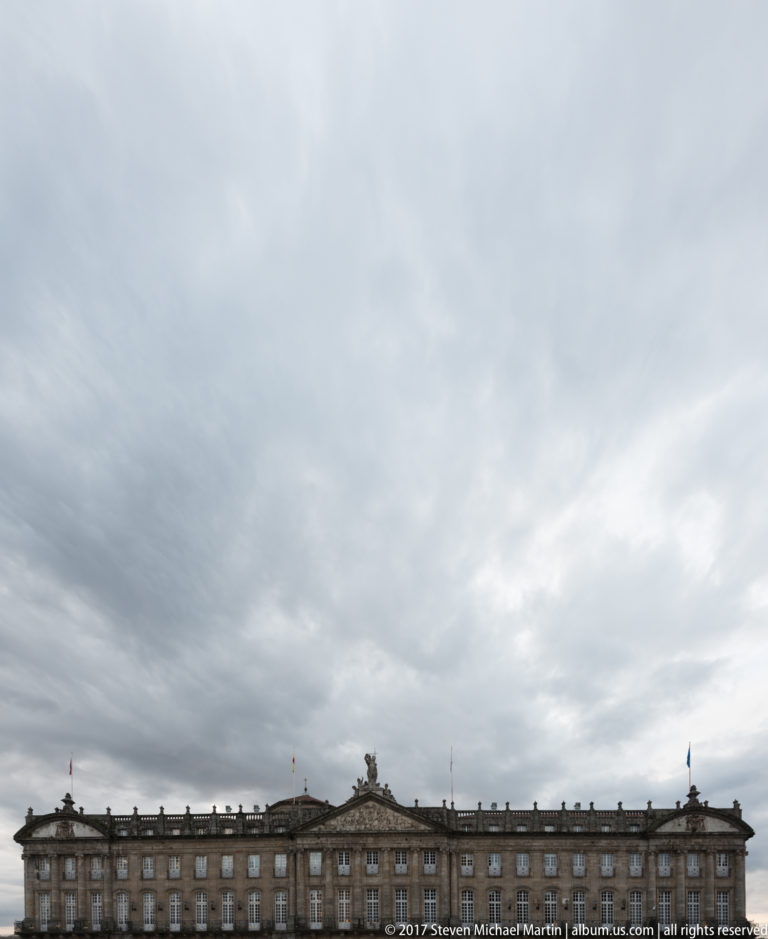 Wide view of Pazo de Raxoi as a low horizontal façade beneath a large overcast sky.
