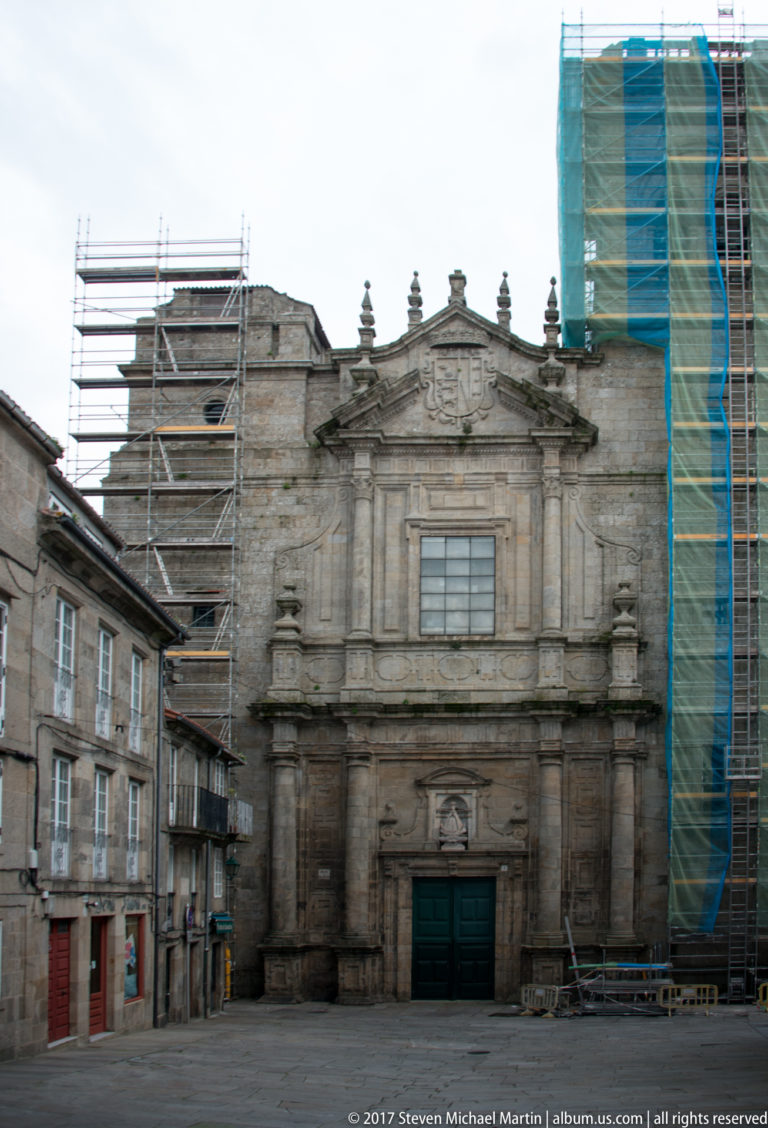 Stone church façade with central doorway and upper window, flanked by old town buildings and restoration scaffolding.