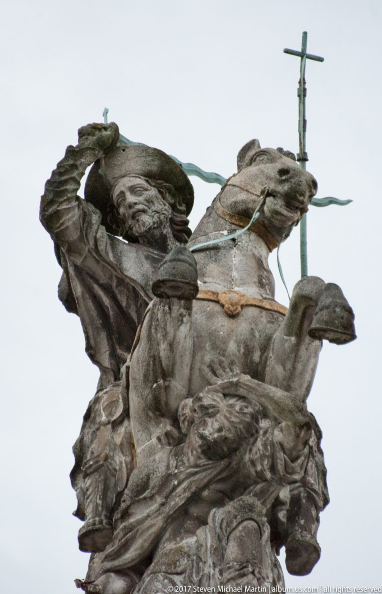Low-angle close view of a weathered bronze sculptural group with mounted and standing figures and a cross-topped staff.