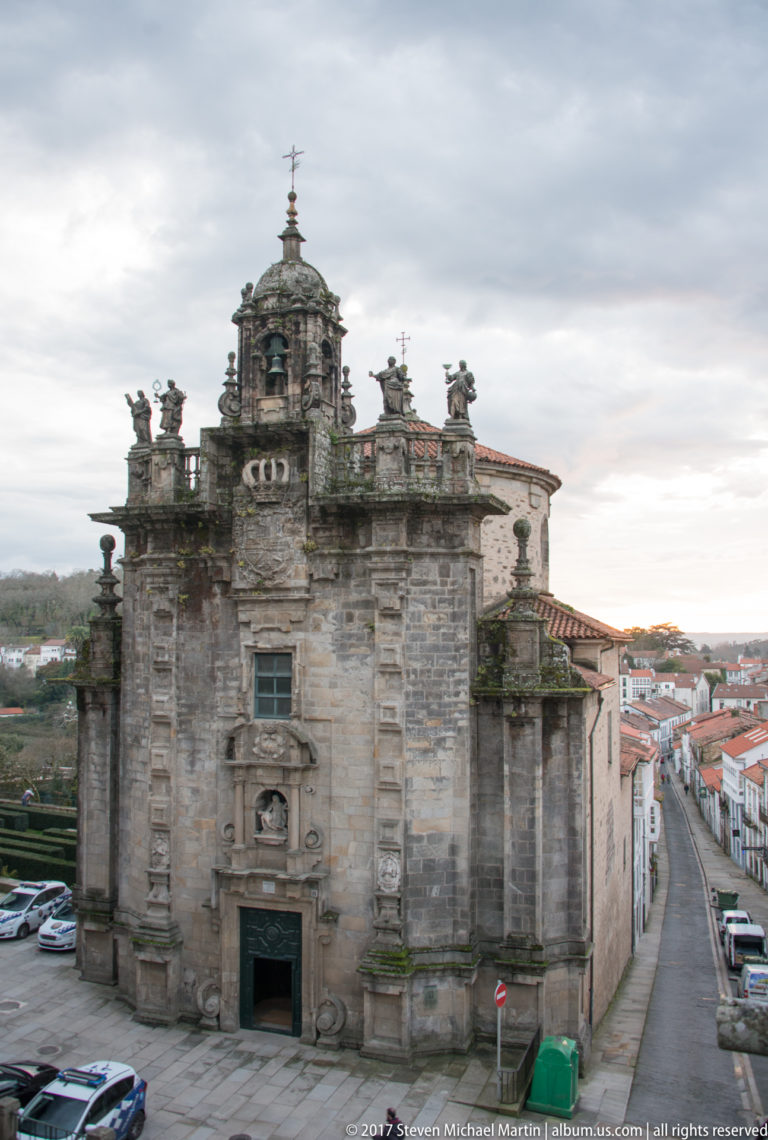 Granite church with central domed bell tower, rooftop statues, and a sloping street beside it.