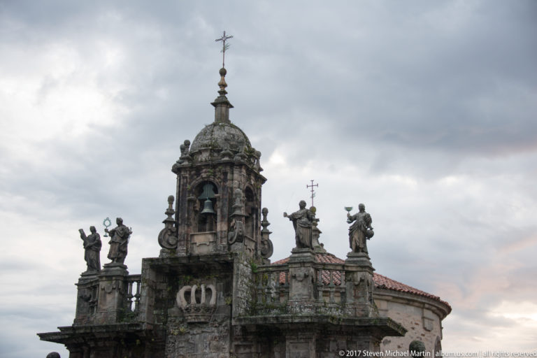 Close view of a granite church bell tower with domed top and statues lining the rooftop parapet.