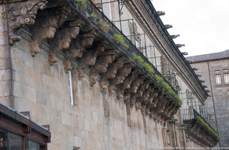 Long iron balcony on a granite building supported by a dense row of carved stone corbels.
