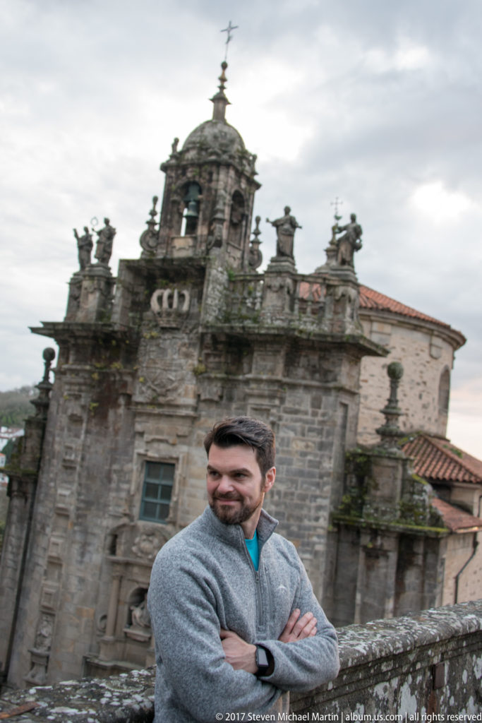 Man in gray sweater standing with arms crossed before a weathered granite church and domed bell tower.