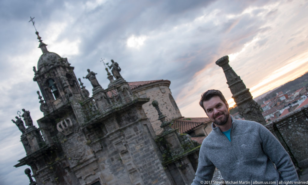 Man in gray sweater beside a stone parapet, with a domed church tower and cloudy sunset sky behind.
